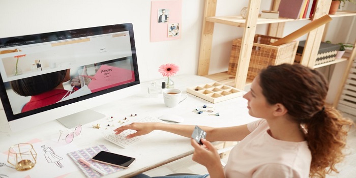 A girl is sitting in front of the computer system, having a card on her hand and accessing the calculator near her, the table holds tea cup, flower and a tray.